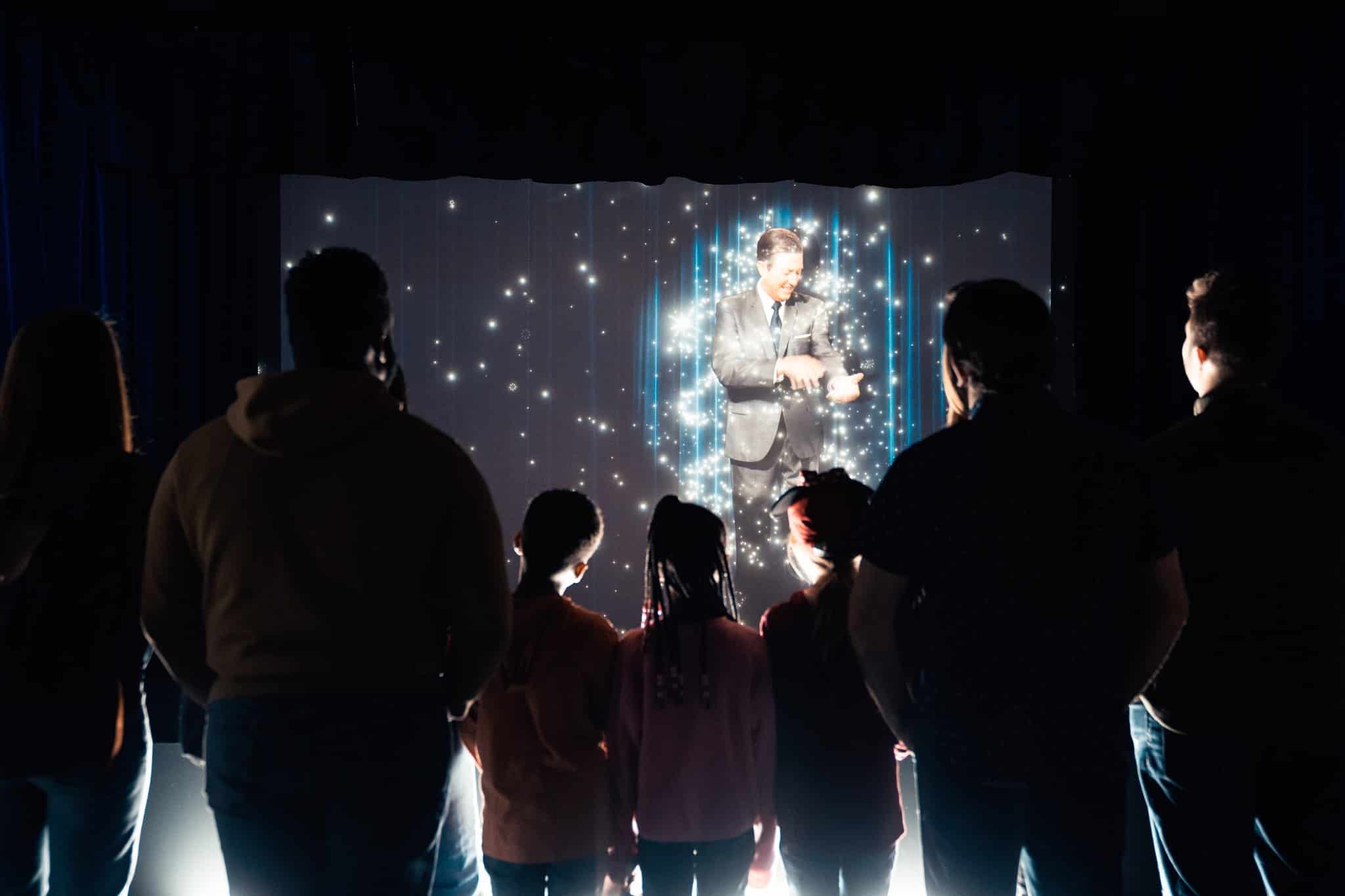 Visitors watch a large movie screen which is playing an introductory video about Walt Disney and his legacy, before walking through the rest of the exhibition.