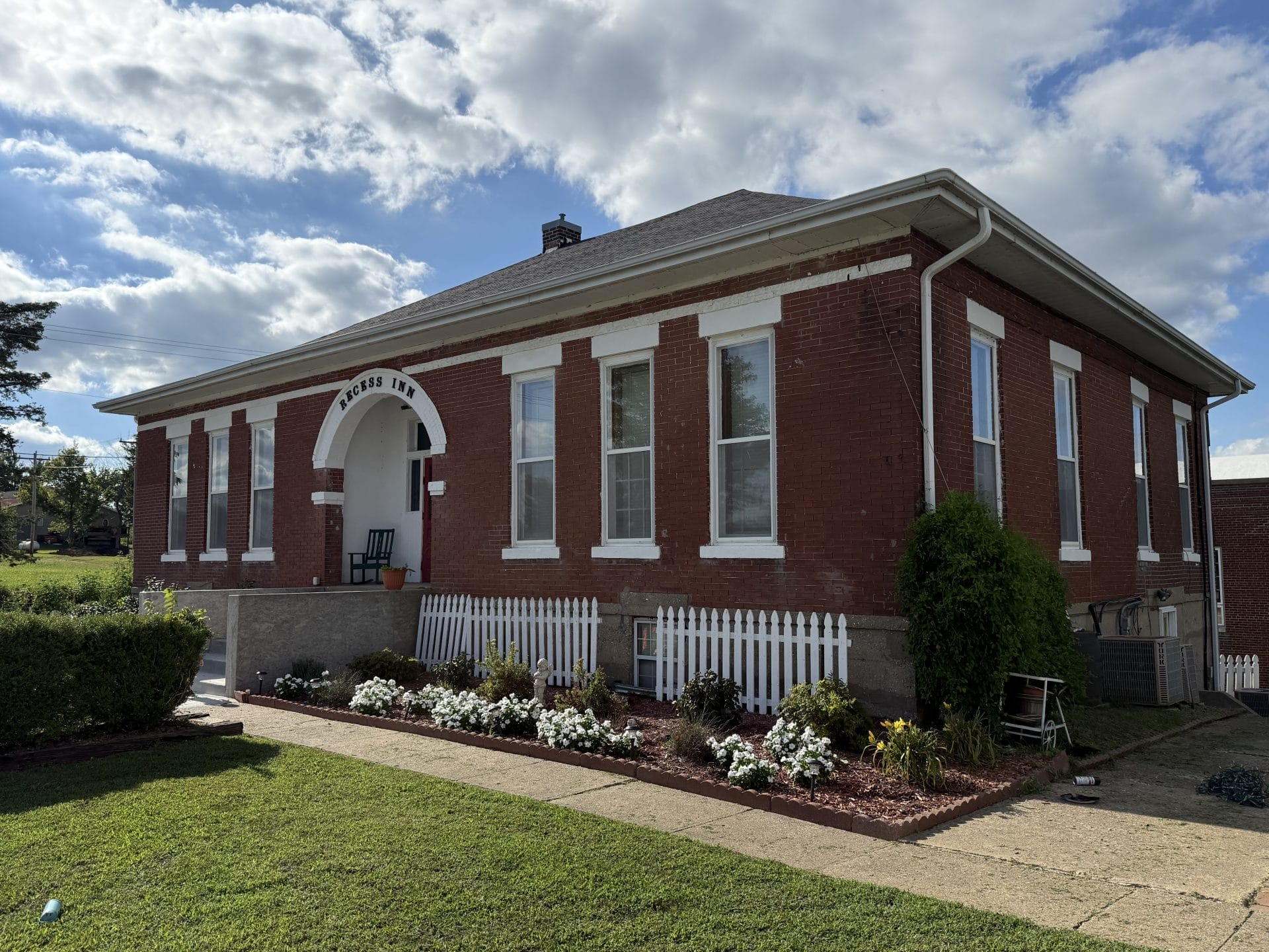 A red brick building with tall windows and a white archway labeled "PARKERS 1915." There is a white picket fence, neatly trimmed shrubs, and a flower bed in front. The sky is partly cloudy.
